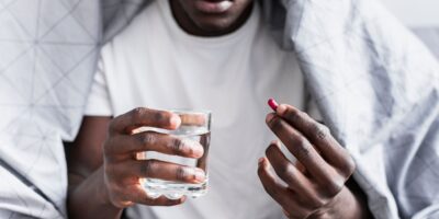 Un jeune homme assis, tenant un verre d’eau dans une main et un comprimé dans l’autre.