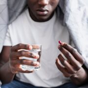 Un jeune homme assis, tenant un verre d’eau dans une main et un comprimé dans l’autre.