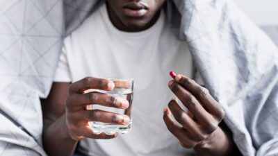 Un jeune homme assis, tenant un verre d’eau dans une main et un comprimé dans l’autre.