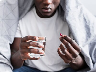 Un jeune homme assis, tenant un verre d’eau dans une main et un comprimé dans l’autre.