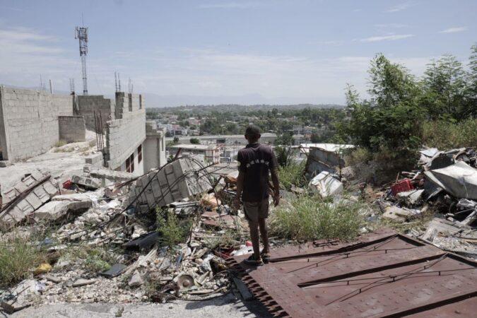 Un homme debout sur une barrière métallique renversée, observant des maisons en ruine à Nazon, le XY août 2025. Photo : Jean Feguens Regala / AyiboPost
